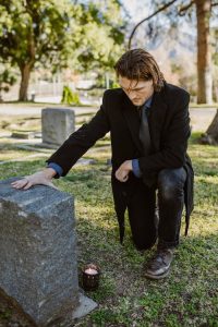A man kneels by a gravestone in a sunny cemetery, reflecting on loss and healing.